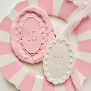Può includere: Un piatto a righe rosa e bianche con bordo smerlato contiene uno stampo in argilla rosa con il numero 10 e un'etichetta bianca con il numero 01. Un nastro rosa con perle è drappeggiato sul piatto.