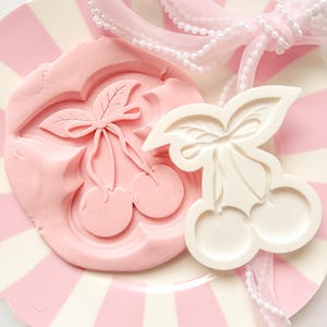 Puede incluir: Un plato a rayas rosas y blancas contiene un molde rosa con forma de cereza y un cortador blanco con forma de cereza. Una cinta rosa con detalles de perlas está drapeada sobre el plato. El molde tiene un diseño detallado de cereza.