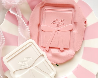 Pode incluir: Um prato listrado rosa e branco contém massa de biscoito rosa com uma impressão em forma de livro e a palavra "Baby" escrita nela. Um cortador de biscoito branco com o mesmo design está ao lado. Uma fita rosa com pérolas está ao fundo.