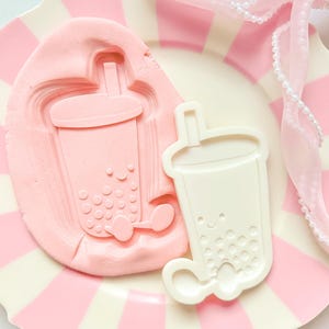 Puede incluir: Un plato a rayas rosas y blancas sostiene un molde rosa y un cortador de galletas blanco, ambos con forma de taza de té de burbujas. Una cinta rosa con perlas está drapeada sobre el plato. El molde tiene una impresión detallada de la taza.