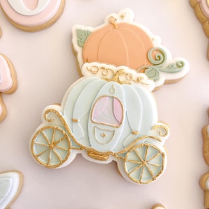Puede incluir: Una galleta con forma de carroza de calabaza azul claro con detalles dorados, que presenta una ventana con cortinas rosas. Detrás hay una galleta separada con forma de calabaza color melocotón con hojas y enredaderas verdes.
