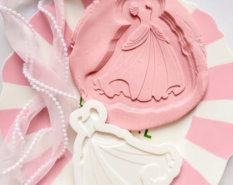 May include: A pink and white striped plate holds a pink clay impression of a dress and a white dress-shaped cutter. A sheer pink ribbon with pearl-like beads is tied in a bow. The items are arranged for a craft project.