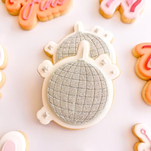Op de afbeelding: Een suikerkoekje in de vorm van een discobal met zilveren glitterglazuur en een witte rand met kleine zilveren steraccenten. Het koekje is omringd door andere versierde suikerkoekjes, waaronder een met de tekst Twenty Fine in oranje en roze.