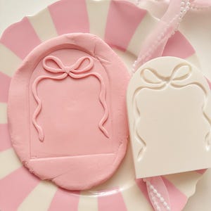 May include: A pink clay mold featuring a bow design, alongside a white mold with the same pattern. They rest on a pink and white striped plate, with a pink ribbon and pearl string. The image suggests a craft or baking theme.