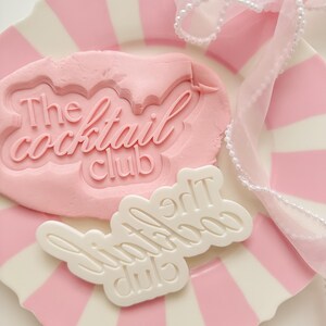 Op de afbeelding: Een roze en wit gestreepte plaat met een roze kleivorm met de tekst "The Cocktail Club". Een witte plastic mal met dezelfde tekst ligt naast de klei. Een doorschijnend roze lint met witte parels is over de plaat gedrapeerd.