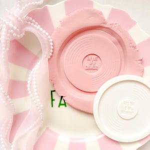 May include: A pink and white striped plate with a pink clay disc that has the text "THE LIFE OF A SHOWGIRL". A white disc with the same text is next to the clay disc. A pink ribbon with white beads is tied around the plate.