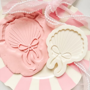 Op de afbeelding: Een roze schelpvormige stempel en de afdruk ervan in roze klei, naast een witte schelpvormige mal. De items liggen op een bord met roze en witte strepen, versierd met een roze lint en een parelsnoer. Het ontwerp heeft een strik.