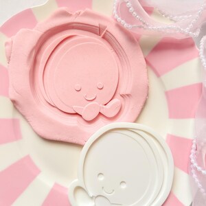 Peut inclure: Une assiette à rayures roses et blanches contient un moule en argile rose et un moule en plastique blanc, tous deux représentant un visage de bébé souriant. Un ruban transparent perlé est drapé sur le côté. Les moules sont probablement pour l'artisanat ou la pâtisserie.