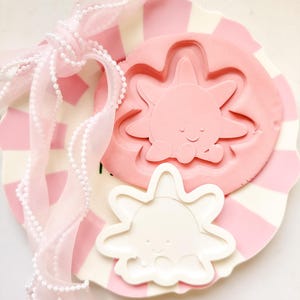 Peut inclure: Une assiette décorative à rayures roses et blanches avec un moule rose en forme de soleil et un moule blanc en forme de soleil. Un ruban rose transparent avec des perles blanches est noué autour de l'assiette. Les moules ont un visage de soleil souriant.