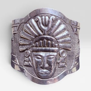 Könnte beinhalten: Ein breiter Armreif aus Silber mit einem zentralen geprägten Relief eines menschlichen Gesichts, das einen aufwendigen Kopfschmuck trägt. Der Hintergrund des zentralen Motivs ist texturiert und punktiert, während die Ränder glatt und poliert sind.