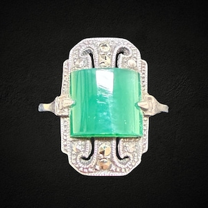May include: A vintage silver ring featuring a rectangular, emerald-green stone. The ring has an ornate, Art Deco design with small, round accents. The ring's band extends to the sides with decorative elements.