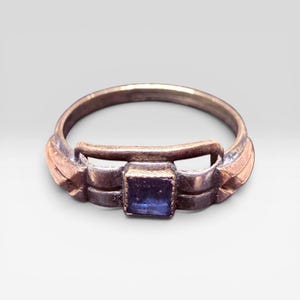 May include: A metallic ring featuring a central square-cut blue stone set within a decorative band. The band has a polished finish with geometric side detailing that frames the stone, creating a structured appearance around the finger loop.