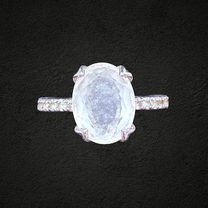 May include: A silver-toned ring featuring a large, oval-shaped, translucent white gemstone held in place by four prongs. The band of the ring is thin and displays a textured, beaded, or pavé-style surface along its outer edges.