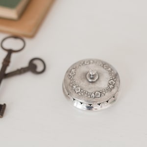 May include: A small, round, silver trinket box with a floral design around the lid's edge and a small knob on top. Two antique keys are crossed in the background, suggesting a vintage or antique theme.