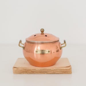 Peut inclure: Une casserole en cuivre avec un couvercle et des poignées en laiton, posée sur une pile de papier vieilli. La casserole a une petite plaque en laiton et un bouton sur le couvercle. La casserole est de couleur brun-rougeâtre.