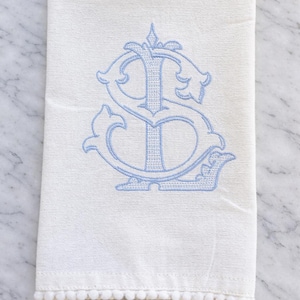 Pode incluir: Toalha de mão de linho branco com monograma bordado em azul claro. O monograma apresenta as letras "S" e "D" num design ornamentado e estilizado. A borda inferior é adornada com pequenos pompons brancos.
