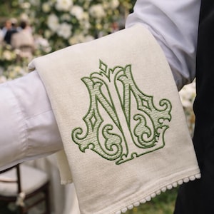 Pode incluir: Guardanapo de linho creme com um monograma bordado em verde. O guardanapo tem uma borda decorativa de pequenos pompons brancos. O monograma é um design ornamentado, provavelmente para um casamento ou evento formal.
