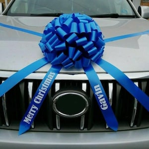 May include: A silver car adorned with a large blue bow on the bonnet. Blue ribbons with the text "Merry Christmas" and "Gavin" are attached. The car's front grill is in view, with the bow as the focal point.