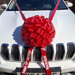 May include: A large red decorative gift bow with two long ribbon tails attached to the bonnet of a white car. The left ribbon tail features the white printed text "Happy 18th Birthday" and the right ribbon tail features the white text "Mackenzie".
