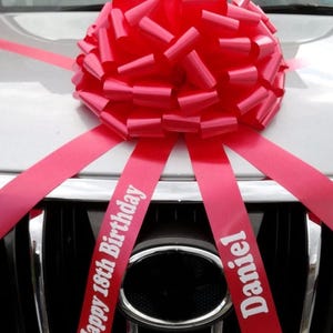 May include: A vibrant red bow, made of layered ribbons, is attached to the front of a silver car. The bow sits above the car's grill. Two ribbons with the text "Happy 18th Birthday Daniel" are draped over the grill, adding a celebratory touch.