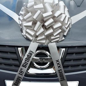 May include: A large silver bow, made of looped ribbon, is attached to the front of a grey car. Two ribbons extend from the bow, one with the text "Happy 18th Birthday" and the other with "James XXX".