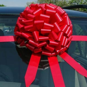 May include: A large, vibrant red bow, made up of many loops, is fixed to the roof of a black car. Long, red ribbons, matching the bow, are used to secure it, adding a celebratory touch to the vehicle's appearance.