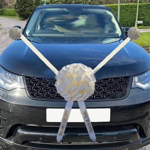 May include: A black car decorated with white bows and ribbons. The ribbons display the text "February 14th 2018" and "Laura & Tom". Smaller bows are attached to the side mirrors. A celebratory decoration for a wedding or event.