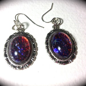 May include: A pair of silver-toned dangle earrings. Each earring features an oval-shaped, iridescent stone with a dark background and flecks of red and blue. The stones are set in a decorative, scalloped frame.