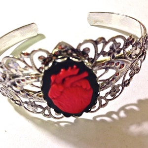 May include: A silver-toned metal cuff bracelet featuring an intricate filigree design with floral motifs. The center of the bracelet displays an oval black cameo setting containing a raised, detailed red anatomical heart sculpture.