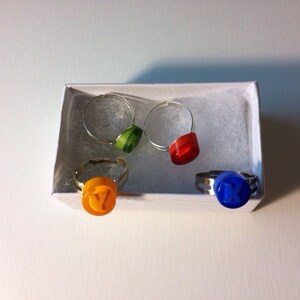 May include: Four adjustable metal rings with circular tops in green, red, orange, and blue. The orange ring features the letter Y, and the blue ring features the letter X. The rings are arranged inside a small white rectangular box with white padding.