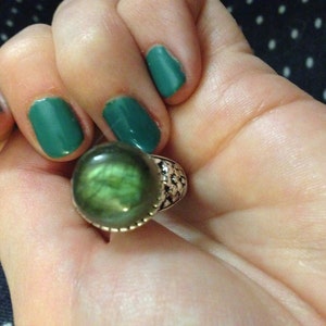 May include: A silver ring featuring a large, round, cabochon-cut labradorite gemstone with a dark green and grey iridescent sheen. The metal band displays a textured, hammered finish around the setting that holds the stone securely in place.