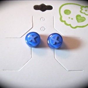 May include: A pair of round, translucent blue stud earrings featuring a raised letter X on the surface. The earrings are attached to a white display card that features a light green graphic of a skull with a flower on its head.