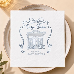 May include: A square white paper napkin featuring a blue line-art illustration of a cafe storefront. The text reads Cafe Bebe, Baby Elliott, and Olivia's Baby Shower. The design is framed by a decorative blue ribbon bow at the top and sides.