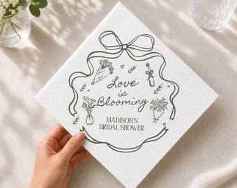 May include: A square white paper napkin featuring a dark green line-art illustration of a ribbon border, floral bouquets, and a bottle. The text reads "Love is Blooming" and "MADISON'S BRIDAL SHOWER" in a dark green script and serif font combination.