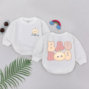 May include: Two white baby bodysuits. One features a dumpling graphic with the name "Anthony" below. The other has "BAO BOO" in pink bubble letters with a dumpling graphic. A rainbow and sunglasses are also in the image.