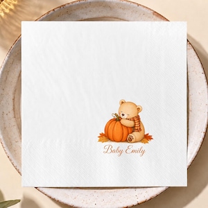 Pode incluir: Um guardanapo de papel branco quadrado com uma ilustração impressa de um urso de peluche castanho claro com um cachecol xadrez a abraçar uma grande abóbora laranja, rodeado por folhas de outono, com o texto Baby Emily em castanho.
