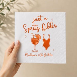 May include: A white square paper napkin featuring orange printed text that reads just a Spritz Older and Maddison's 40th Birthday. The design includes an orange illustration of a cocktail in a stemmed glass with a citrus slice and a one-piece swimsuit.