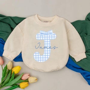 May include: Cream-colored baby romper with long sleeves. Features a light blue gingham "J" with a bow and the name "James" in cursive. The romper is made of soft material, perfect for a baby.