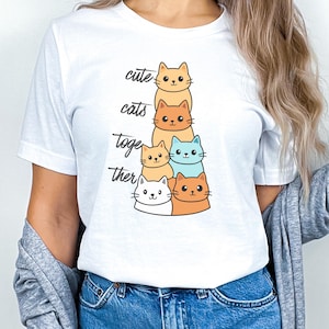 Puede incluir: Camiseta blanca con un diseño de gatos de dibujos animados apilados en tonos naranja, amarillo y azul. Las palabras "cute cats together" están impresas verticalmente en la camiseta con una fuente lúdica. La camiseta está hecha de una tela suave.