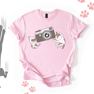 Peut inclure: T-shirt rose clair avec un dessin animé de deux chats tenant un appareil photo marron. Les chats sont blancs et bruns. Des empreintes de pattes sont dispersées.