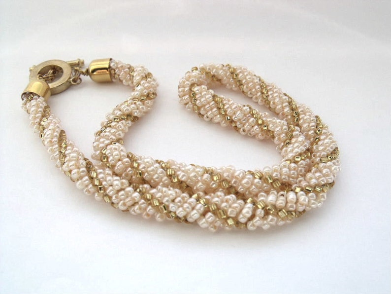 May include: A multi-strand necklace made of small white and gold beads woven together in a twisted pattern. The necklace has a gold clasp.