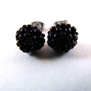 May include: A pair of black stud earrings. Each earring is a sphere covered in small, round, glossy black beads. The posts are silver-colored. The earrings are set against a white background.