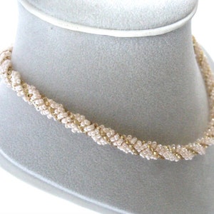 May include: A delicate gold and ivory beaded necklace with a twisted design. The necklace is displayed on a white mannequin.