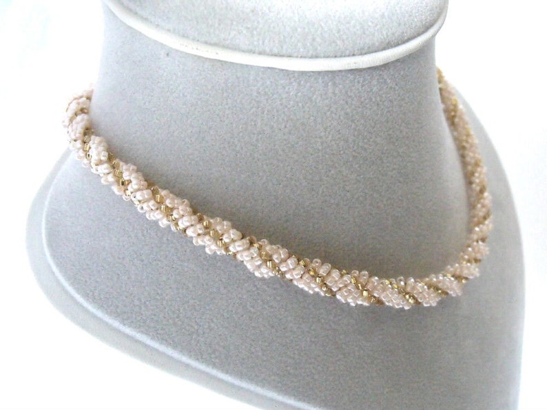 May include: A delicate gold and ivory beaded necklace with a twisted design. The necklace is displayed on a white mannequin.