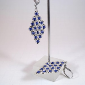 May include: A pair of diamond-shaped earrings with a silver-colored frame and dark blue beads. The earrings are displayed on a white surface, with one earring hanging and the other resting on the surface. The earrings have a lever back closure.