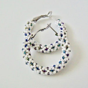 May include: A pair of white beaded hoop earrings with silver-tone clasps. The earrings are adorned with small, colorful beads in shades of blue, green, and purple, creating a textured surface. The hoops are approximately 1.5 inches in diameter.