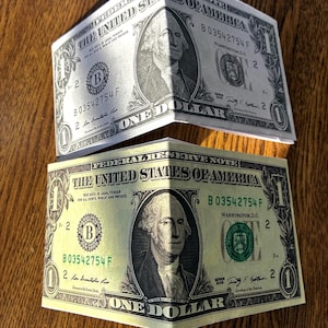 Puede incluir: Dos billetes de un dólar estadounidense doblados por la mitad horizontalmente. Cada billete presenta el retrato de George Washington en el centro, el texto "The United States of America" en la parte superior y "One Dollar" en la parte inferior.