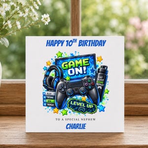 Peut inclure: Une carte d'anniversaire carrée avec le texte HAPPY 10TH BIRTHDAY, TO A SPECIAL NEPHEW et CHARLIE. L'illustration montre une manette de jeu noire, un casque bleu, une gourde avec EAT SLEEP GAME REPEAT et les mots GAME ON! LEVEL UP.