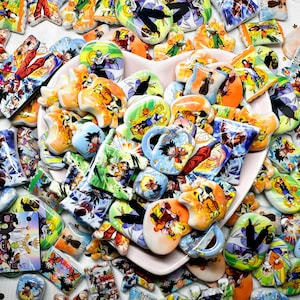 May include: A collection of colorful ceramic pieces featuring various characters and scenes from a popular anime series. The pieces are in various shapes, including hearts, rectangles, and mugs, with vibrant orange, blue, and green hues.