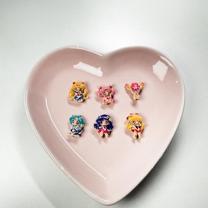 Może przedstawiać: Sześć miniaturowych figurek postaci z anime, każda z unikalnymi, kolorowymi strojami i fryzurami, ułożonych na różowym talerzu w kształcie serca. Figurki mają detale takie jak kokardy, wstążki i akcesoria, na białym tle.